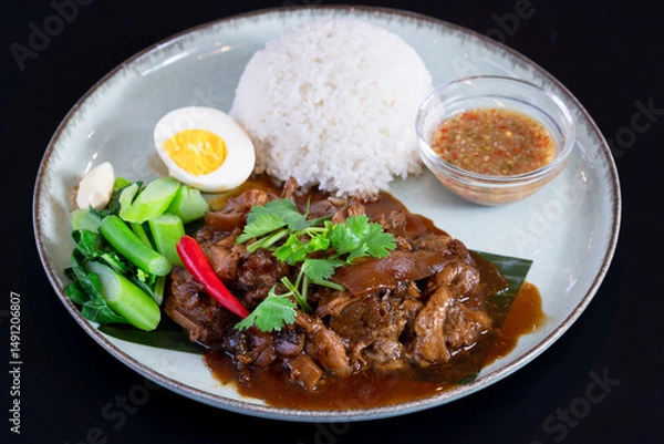 Fototapeta A plate of Asian braised stew slow cooked pork with white rice with its spicy sauce with garlic, topped with green coriander black isolated background.