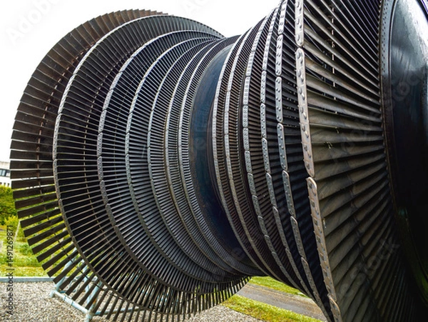 Fototapeta Massive industrial turbine rotor with closely spaced, curved metal blades arranged in concentric rows, displayed outdoors on a gravel platform in a grassy area.