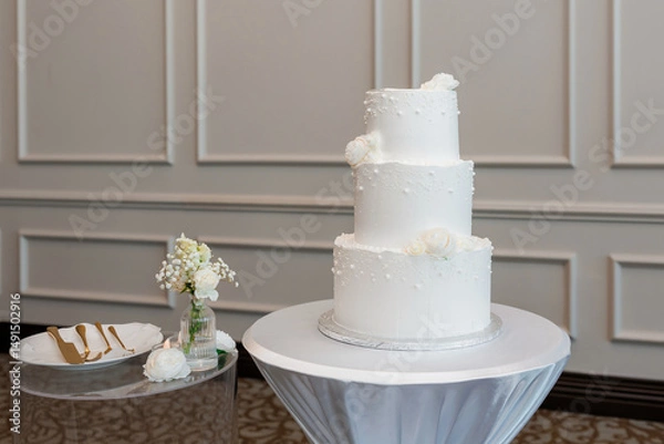 Obraz Multi-tiered wedding cake covered with white vanilla cream. Three-tiered wedding cake stands on a table in a restaurant ready to be cut by the newlyweds. Dessert for guests at a wedding banquet