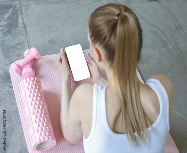 Fototapeta Woman lying on yoga mat after workout, holding smartphone with blank screen. Pink dumbbells and foam roller nearby. Concept of fitness tracking and mobile app use.