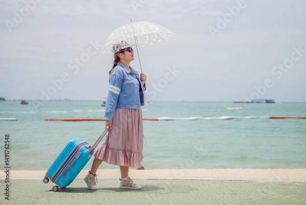 Fototapeta Woman Traveler walking with Luggage and Umbrella by the Seaside. A female tourist with a blue suitcase and an umbrella enjoys a sunny day by the sea, strolling along the waterfront.