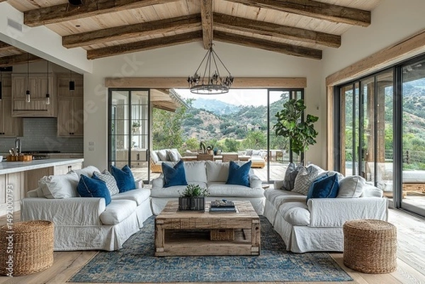 Fototapeta Modern farmhouse living room with white walls, light wood floors and ceiling beams, blue accent pillows, large chandelier, open kitchen, and glass sliding doors leading to outdoor deck with valley vie