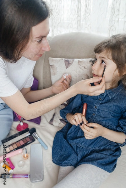 Fototapeta Mother putting makeup on her daughter face and they fool around together. Communication between women in the family. Fun time. Happy family concept. little girl doing her makeup