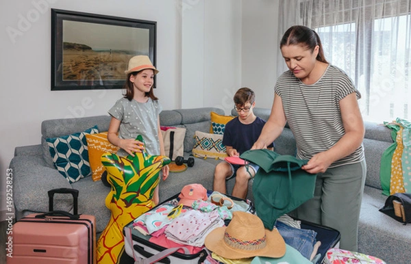 Fototapeta Family packing suitcases for summer holidays in living room. Smiling daughter holding inflatable pineapple float and mother folding swimwear to suitcase trip while son playing with ping pong paddles.
