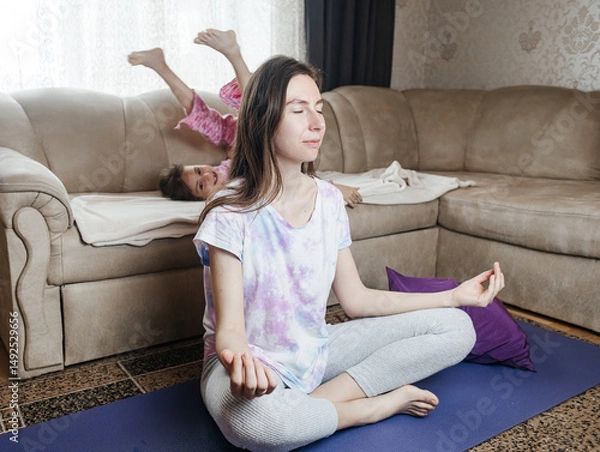 Fototapeta Young mother tries to meditate and do a home workout while her playful daughter gets in her way. Interrupted yoga at home and parenting reality. Mom and daughter having fun
