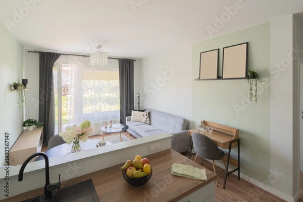 Fototapeta Living and dining salon viewed from a modern kitchen. The open space shows a dining set, sofa, and large windows. Kitchen island and green accent wall frame this bright European home interior.