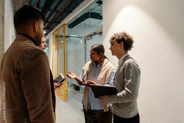 Fototapeta A young White man with a notebook and an Indian adult woman are talking with a white-skinned man with a tablet and a dark-skinned man while standing in an office corridor