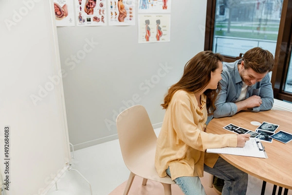 Fototapeta The White adult man and woman sit at a table in a medical office, smiling and reviewing ultrasound images together, with anatomical posters on the wall indicating a prenatal consultation.