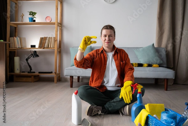 Obraz Housekeeping job concept. Young woman in casual clothes sitting on floor with cleaning supplies, washing and doing homework cleaning routine looking at camera showing small gesture