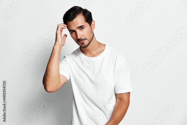 Fototapeta Attractive young man touching hair smiles confidently while posing in a casual white t-shirt against plain light background. Modern lifestyle concept.