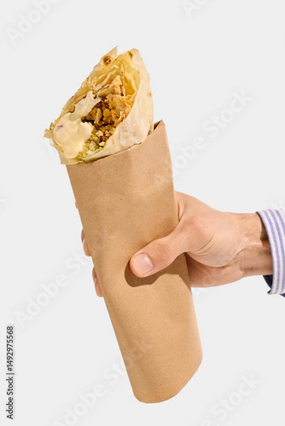 Fototapeta Shawarma roll with grilled meat, white sauce, cabbage, and flatbread held in brown paper sleeve, viewed from hand against white background. Concept of shawarma, wrap, meat, takeaway, street food