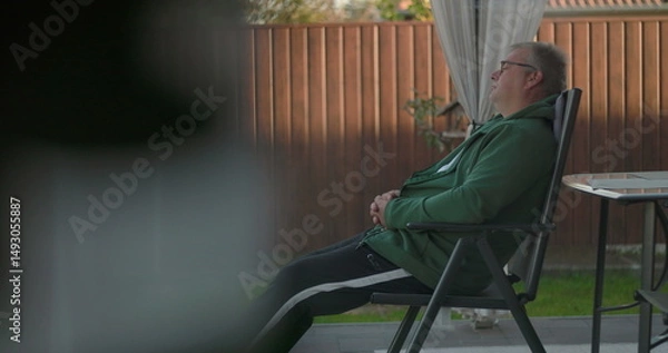 Fototapeta Man reclining on patio chair with arms crossed, resting in backyard with wooden fence and greenery, enjoying quiet relaxation and outdoor serenity