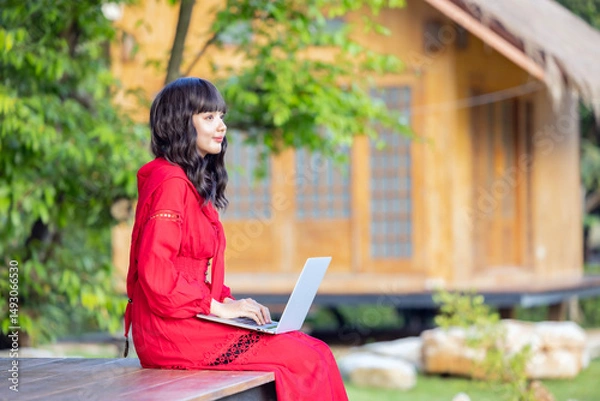 Obraz Side view of Asian woman smiling happily while sitting on cottage terrace in the countryside with laptop working remotely on new web design project in sunny garden.