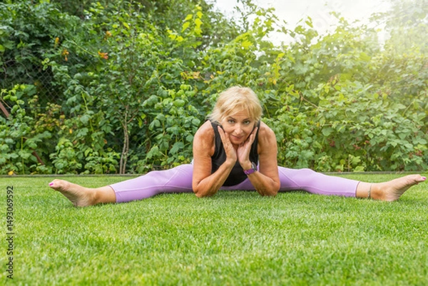 Obraz Flexible smiling mature woman  doing a leg split supporting her chin with both hands. Horizontally. 