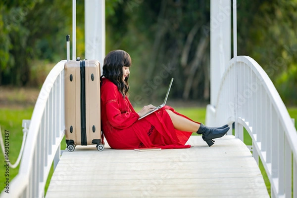 Obraz Young woman wear summer casual clothes sit near suitcase using laptop for online search for holiday destination or discount offers on travel while sitting on bridge relaxing outdoors feeling happiness