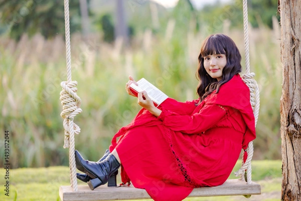 Obraz Beautiful Asian woman feeling happy smiling and looking to camera while relax sitting on a swing at flowering field and reading book.