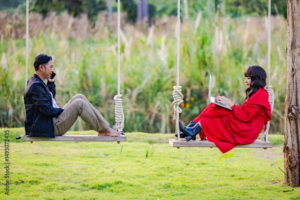 Obraz Weekends getaway. Asian couple enjoying the joys of being on an outdoor wooden swing during sunset, searching sightseeings for trip on laptop, planning route and talking on cellphone.