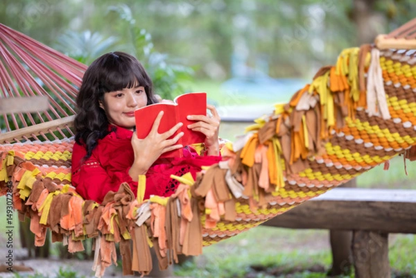 Obraz Young elegant beautiful Asian woman dressed in summer clothes smiling and  reading book lying on hammock at terrace.