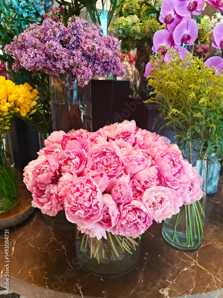 Fototapeta A bouquet of pink peonies sits in a clear vase, surrounded by a vibrant display of other flowers including orchids and lilacs. The setting is a lively floral shop.