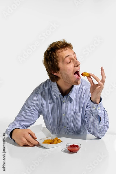 Fototapeta Young man about to eat crispy chicken nugget with wide open mouth and dipping sauce on table, enjoying quick meal against white background. Concept of fast food, catering, nutrition, traditional.