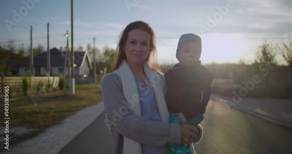 Fototapeta Mother carrying toddler in arms while smiling outdoors on a quiet suburban street, enjoying a peaceful moment during a sunny day near neighborhood homes