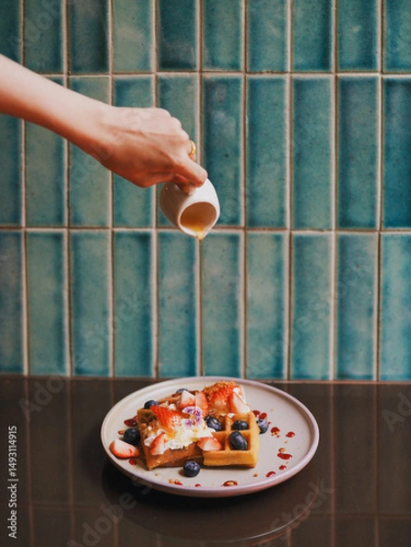 Fototapeta Waffles with fresh berries. A hand pouring maple syrup on waffles with topping whipping cream and berries (strawberries and blueberries)