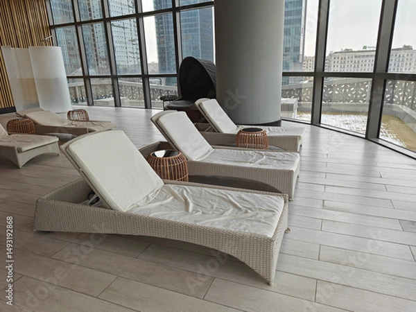 Fototapeta Contemporary lounge space with wicker sun loungers and soft cushions, large windows revealing city skyline, inviting relaxation and comfort in a tranquil atmosphere