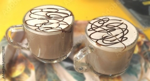 Fototapeta Two glass cups of hot chocolate with frothy milk and artistic chocolate syrup topping, served on a colorful tray with a vibrant background.