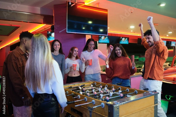 Fototapeta Group of friends having fun playing table football and cheering together in a colorful amusement arcade