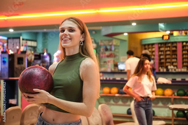 Obraz Happy young woman holding bowling ball, smiling in bowling alley, ready for a game with friends