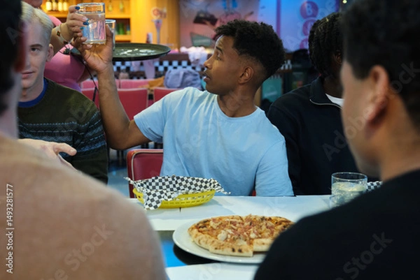 Obraz Friends enjoying pizza and drinks in a diner, a young man taking a glass of water from the waiter