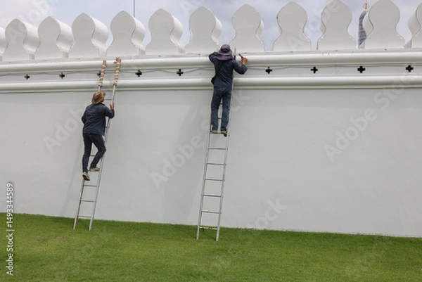 Fototapeta Workers are painting a large white wall with ladders in a beautiful outdoor setting.