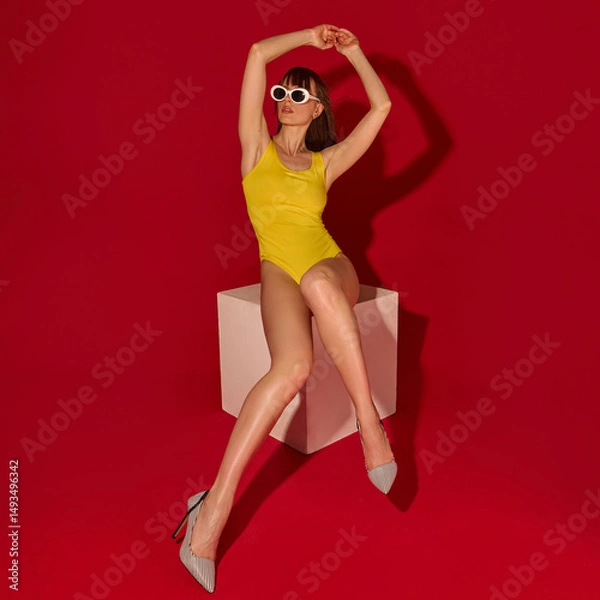 Fototapeta Beautiful female model in yellow swimsuit stretches arms up while sitting on white cube showing legs style and summer fashion against red background. Concept of beauty, fashion, style.