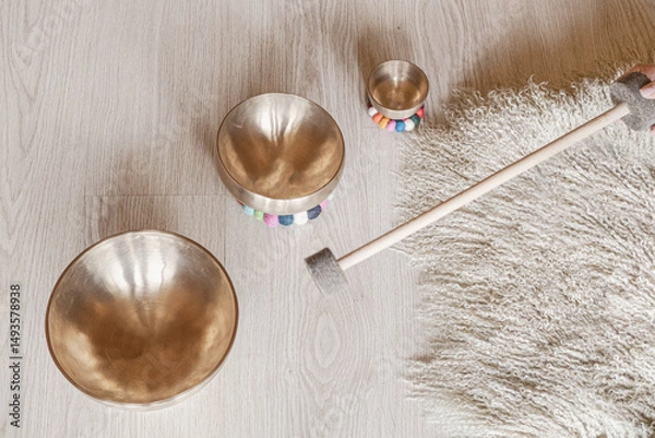Obraz Tibetan singing bowls of varying sizes on a light wooden floor with a fluffy rug. A hand holding a mallet approaches the bowls, suggesting an impending sound therapy session