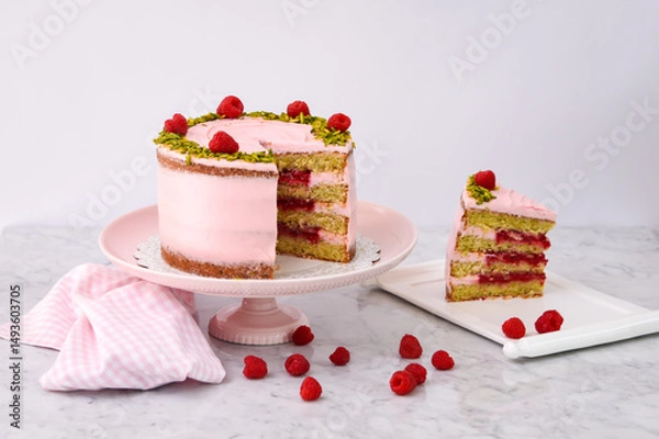 Fototapeta ayered Raspberry Pistachio Cake with a slice ith light pink cream and garnished with fresh raspberries and chopped pistachios side view of delicious dessert