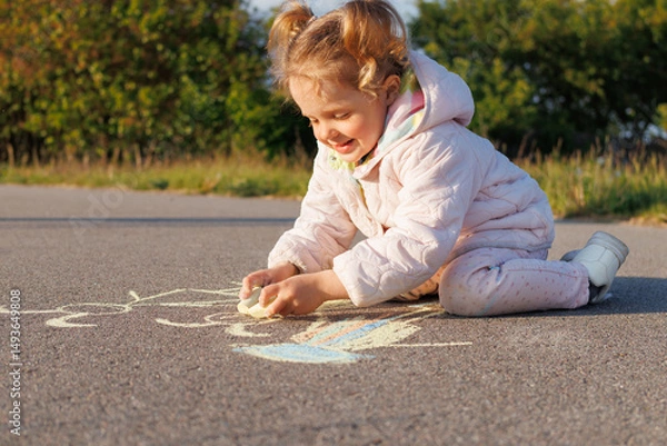 Fototapeta A smiling toddler in a pink jacket draws with white chalk on asphalt, enjoying outdoor play on a sunny day.