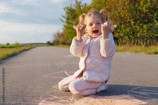 Fototapeta A joyous toddler in a pink jacket excitedly holds chalk, looking up and laughing while sitting on asphalt with drawings around.