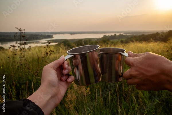 Obraz Two hands holding stainless steel camping mugs in a toast against a scenic sunset landscape with rolling hills and a winding river symbolizing outdoor adventure travel and shared moments in nature.
