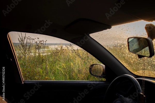 Obraz Looking out from inside a car through a window covered in raindrops reveals a hazy landscape of vegetation and a body of water under a soft evening light suggesting a journey on a wet road.