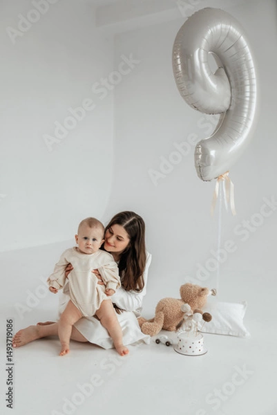 Fototapeta Young mother sitting with her 9 month old son on the floor on a white background with a balloon number 9