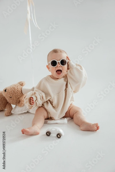 Fototapeta Handsome little boy in a beige sweater with toys and glasses sits on the floor and laughs. Children's emotions and laughter