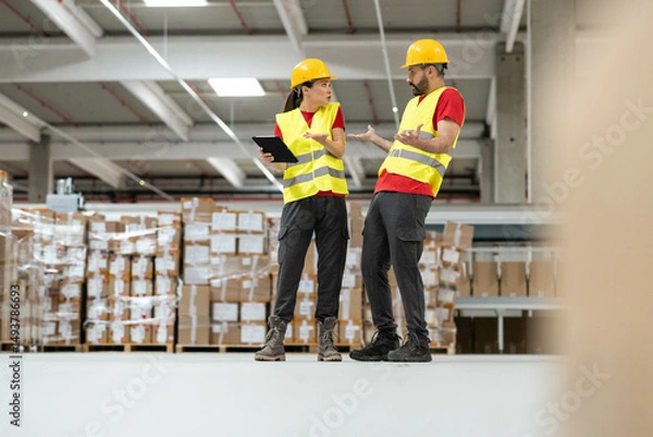 Fototapeta Two warehouse workers engage in a lively discussion while using a tablet in a bustling storage environment filled with boxes.
