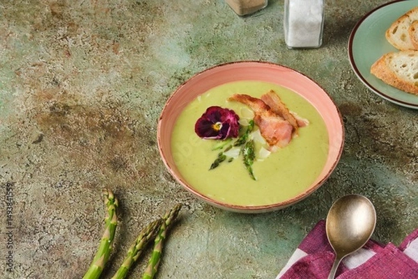 Fototapeta Fresh asparagus puree soup with asparagus pieces and fried bacon in a pink ceramic plate on a green concrete background. Recipes asparagus.