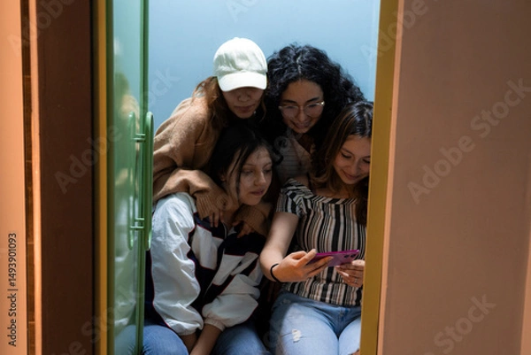 Obraz Four cheerful friends gather around a smartphone, sharing joyful and amusing moments