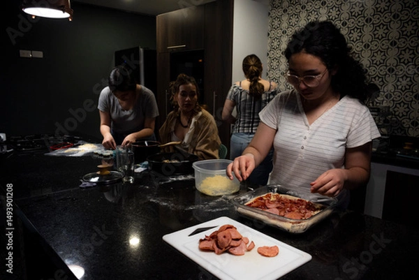 Obraz Four women collaborate in a kitchen, making pizza, food using dough and molds, capturing moments of teamwork and friendship in a warm, homey environment for an enjoyable culinary experience.