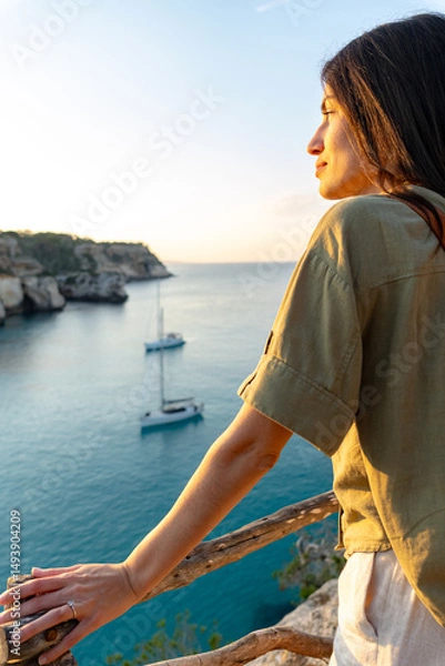 Fototapeta Woman standing on a balcony overlooking a tranquil ocean scene with sailboats, enjoying the serene sunset and natural beauty