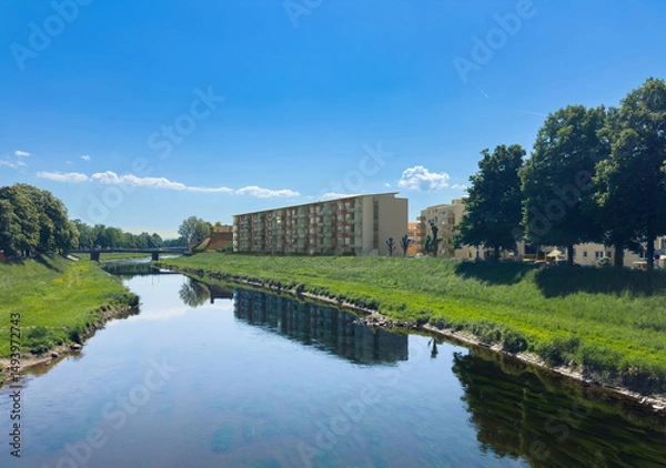 Fototapeta Scenic view of the Murg river in Rastatt, Germany with modern residential buildings on the right bank and lush greenery reflecting in the calm water under a bright blue sky