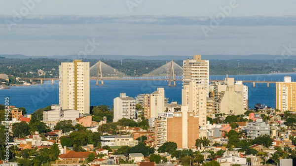 Obraz Panoramic view of the city of Encarnación, Paraguay, with modern buildings in the foreground and the San Roque González international bridge crossing the Paraná River in the background.