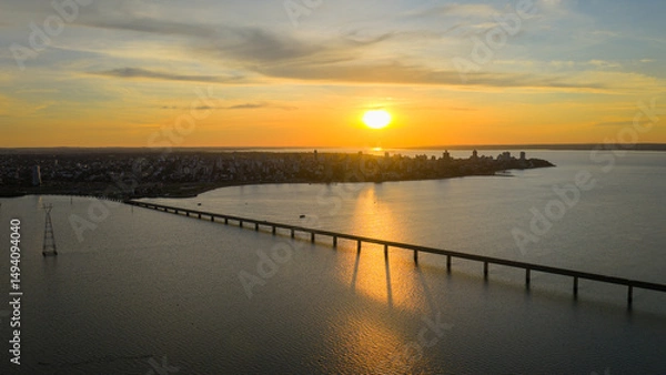 Obraz Golden hour aerial view of Posadas, Misiones, Argentina, with the Paraná River reflecting the sunset and part of the San Roque González bridge visible on the right.