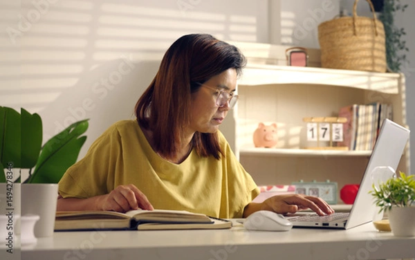 Fototapeta Asian woman works from home, surrounded by notes, a laptop, and messages. She browses the internet, writes ideas, and researches for her startup, utilizing wireless technology.